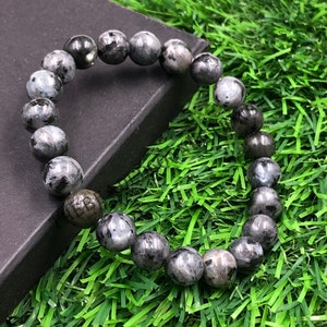 Pode incluir: Uma pulseira de contas redondas de labradorite cinza com inclusões escuras. As contas polidas são enfiadas. A pulseira está sobre uma superfície preta, sobre um fundo de relva verde.