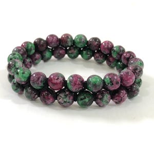 May include: A double-strand bracelet composed of polished, spherical beads featuring a mottled pattern of deep magenta and forest green. The beads are strung together on a thin black cord, creating a layered appearance with a smooth, glossy finish.