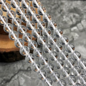 May include: A strand of clear quartz beads, each bead is round and smooth. The beads are strung on a clear thread and are arranged in a row. The beads are all the same size and are evenly spaced.