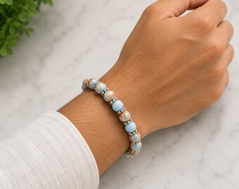 Pode incluir: Uma pulseira de contas com contas redondas de pedra marmoreada em azul claro e bege, alternadas com pequenas contas espaçadoras metálicas em tom prateado. A pulseira é construída com uma série destas contas esféricas num padrão repetitivo.