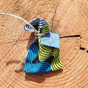 May include: A small, colorful fabric pouch with a blue, yellow, and black pattern. The pouch is tied with twine and has a blue tag attached with the text "Merci de Lunik" written in black.