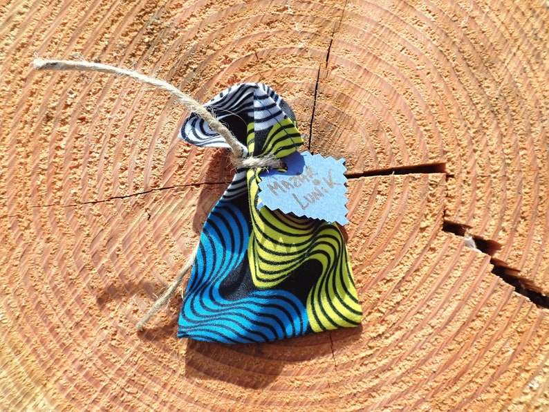 May include: A small, colorful fabric pouch with a blue, yellow, and black pattern. The pouch is tied with twine and has a blue tag attached with the text "Merci de Lunik" written in black.