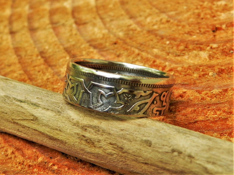 May include: A silver ring made from a repurposed coin with intricate designs and lettering. The ring is resting on a piece of weathered wood.