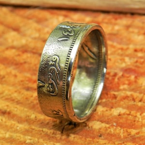 May include: A silver coin ring with Arabic script and a textured surface. The ring is on a wooden surface.