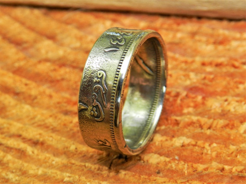 May include: A silver coin ring with Arabic script and a textured surface. The ring is on a wooden surface.