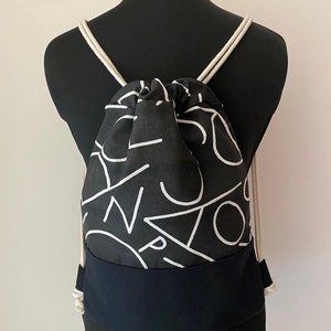 May include: Black drawstring backpack with white geometric patterns and a black bottom panel. The backpack has white drawstrings.
