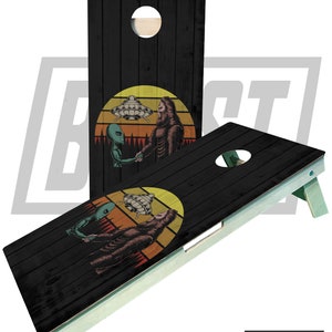 May include: A black wooden cornhole board with a retro sunset design featuring an alien and Bigfoot shaking hands. The alien is green and has a spaceship above it. The Bigfoot is brown and has fur. The background is a sunset with orange, yellow, and red colors.