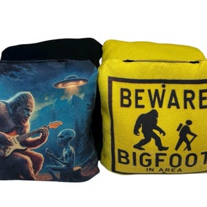 May include: Two yellow and blue bean bags with printed designs. One bean bag features a Bigfoot playing a guitar with an alien. The other bean bag has a yellow warning sign that reads "Beware Bigfoot in Area".