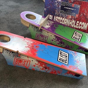 May include: Three wooden cornhole boards with a red, green, and blue paint splatter design. The boards have a hole in the top and the text "BLAST" printed on them. The boards are for the game of cornhole.