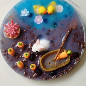 May include: A miniature scene with a white rabbit, two yellow rubber duckies, a small potted plant, and a brown shovel in a basket. The scene is set in a purple and blue resin with brown beads and small orange carrots.
