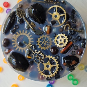 May include: A round, clear resin coaster with a dark blue background. The coaster is filled with various small objects, including black stones, gold gears, keys, and beads.