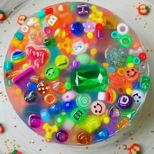 May include: A clear, round resin paperweight filled with colorful beads, letters, and small toys. The paperweight has a large green gem in the center.