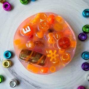 May include: A clear, round, pink resin soap with a variety of colorful beads and shapes embedded inside. The soap is surrounded by colorful beads.