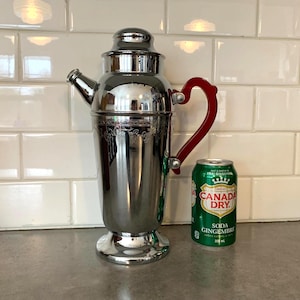 Könnte beinhalten: Ein Vintage-Cocktailshaker aus Chrom mit rotem Griff und Ausguss. Der Shaker hat ein verziertes Design in der Mitte und steht auf einem runden Sockel. Eine grün-weiße Dose Canada Dry Ginger Ale steht daneben.