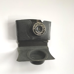 Pode incluir: Câmera Clix Deluxe preta vintage com estojo de couro. A câmera tem uma lente circular e as palavras "CLIX DE LUXE" são impressas na frente. O estojo está aberto, revelando a lente e um compartimento.