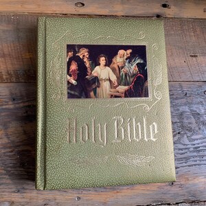 Könnte beinhalten: Ein grünes Hardcover-Buch mit goldenen Lettern, auf dem "Holy Bible" steht. Der Umschlag zeigt eine Vintage-Illustration von Menschen in einem Raum.
