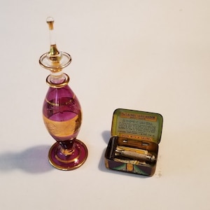 Pode incluir: Uma garrafa de vidro roxo com detalhes dourados e um tampão de vidro transparente, ao lado de uma caixa de lata aberta contendo uma lâmina de barbear de segurança com o texto "The "Salvo" Safety Razor" impresso na caixa.