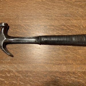 Peut inclure: Un marteau arrache-clous avec une tête en métal foncé et un manche marron foncé texturé. Le marteau est posé sur une surface en bois. Le marteau arrache-clous est un outil utilisé pour la construction et la menuiserie.