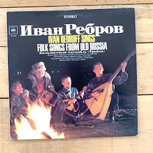 Op de afbeelding: Een vintage vinyl platenhoes met een foto van drie mannen die traditionele Russische instrumenten bespelen rond een kampvuur. De hoes is zwart met witte tekst die "Ivan Rebroff Sings Folk Songs From Old Russia" en "Балалаечный Ансамбль «Тройка»" luidt.