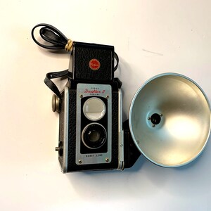 Peut inclure: Un appareil photo Kodak Duaflex II vintage, avec un corps noir, des accents argentés et un grand flash argenté. L'appareil photo est équipé d'une sangle en cuir et d'un viseur noir monté sur le dessus. Le logo Kodak est visible.