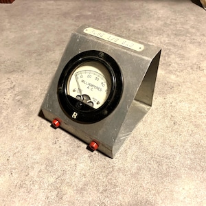 Puede incluir: Un antiguo miliamperímetro plateado con un indicador circular negro. El indicador tiene una esfera blanca con marcas negras y una aguja. El medidor está alojado en una carcasa metálica con terminales rojos y el texto "FUSE L/S BAR".