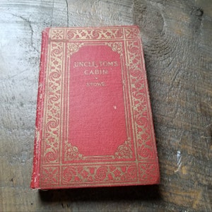 Puede incluir: Una copia antigua de "La cabaña del tío Tom" de Stowe. El libro tiene una cubierta roja con bordes y letras doradas decorativas. El título y el nombre del autor son visibles en la portada. El libro está ligeramente desgastado, lo que sugiere su edad.