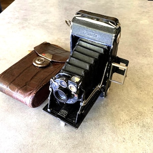 May include: A classic black folding camera with a bellows design, alongside a brown leather case. The camera has a lens and dials, with "Roonta" on top. The case features a metal clasp. A vintage photography item.