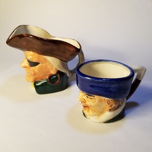 Könnte beinhalten: Zwei Keramikbecher in Form von Männerköpfen. Einer ist ein Krug mit braunem Hut und grüner Jacke. Der andere ist ein Becher mit blauem Hut. Beide haben einen weißen Innenraum und einen Henkel.