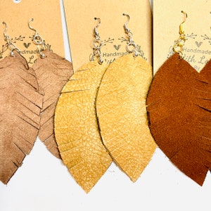 May include: Three pairs of leather earrings with fringe. The earrings are in brown, tan, and gold. The earrings are handmade with love.