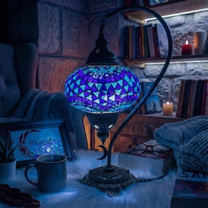 May include: A mosaic glass table lamp in blue and purple hues. The lamp features a curved metal frame and ornate base. The lamp is lit, casting a soft glow. The lamp is on a table with a mug, books, and other decorative items, creating a cozy atmosphere.