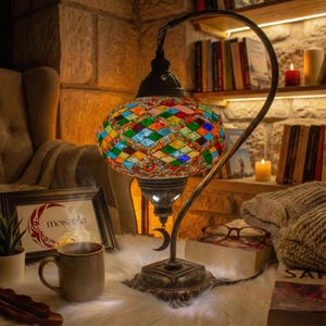 Puede incluir: Lámpara de mesa decorativa con pantalla de cristal de mosaico colorido. La lámpara tiene una base de metal color bronce y un brazo curvo. La pantalla es una mezcla de piezas de vidrio rojas, azules, verdes y amarillas. Detalle de luna.