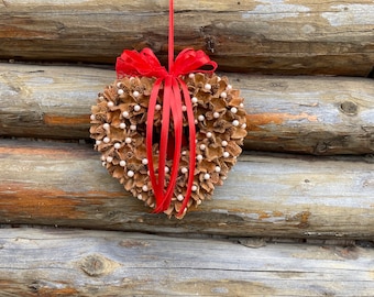 May include: A heart-shaped wreath made of pine cones and white beads with a red ribbon bow.