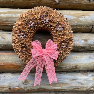 Op de afbeelding: Een bruine dennenappelenkrans met een roze kant strik. De krans hangt aan een houten muur.