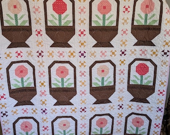 May include: A white quilt with a pink border. The quilt features a repeating pattern of brown flower pots with pink flowers and green leaves. The flower pots are surrounded by a white background with a repeating pattern of small pink, white, and yellow triangles.