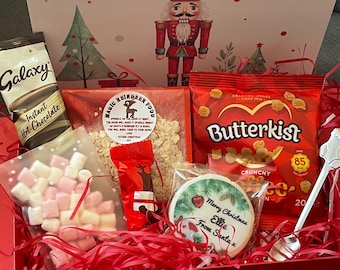 May include: A red Christmas Eve gift box containing Galaxy hot chocolate, marshmallows, Butterkist popcorn, and a decorated cookie. A toy nutcracker is inside, with red paper filler. The box lid says 'Christmas Eve'.