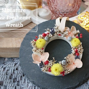 May include: A white circular wreath with dried flowers and berries. The wreath is decorated with yellow, pink, and blue flowers, as well as red berries. The wreath is on a black slate surface. Miniature Air Freshener Wreath.