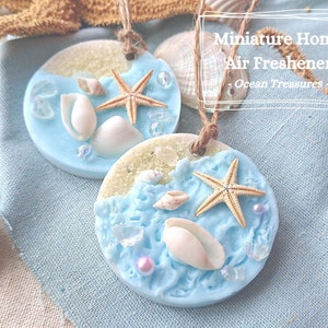 May include: Two round, blue and white air fresheners with a beach theme. Each air freshener features a starfish, seashells, and glitter. The air fresheners are hanging from a twine string. The text "Miniature Home Air Freshener - Ocean Treasures -" is visible in the image.