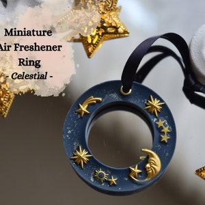 May include: A dark blue, round air freshener ring with a celestial theme. It features gold stars, a crescent moon, and a shooting star. The ring is attached to a black ribbon.