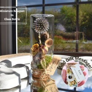 May include: A miniature bouquet of dried flowers and greenery in a clear glass bottle with a cork stopper. The bottle is tied with twine and a small tag that says "Gemini Herbarium".