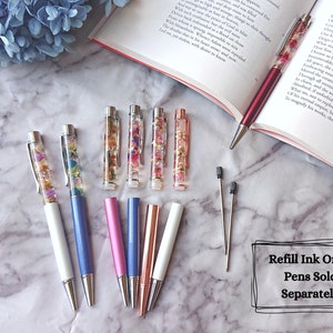 Peut inclure: Un ensemble de huit stylos avec des fleurs color&eacute;es et des paillettes enferm&eacute;es dans de la r&eacute;sine transparente. Les stylos ont des corps de diff&eacute;rentes couleurs, notamment blanc, bleu, rose et or rose. Le texte "Recharge d'encre uniquement ! Stylos vendus s&eacute;par&eacute;ment" est visible sur l'image.