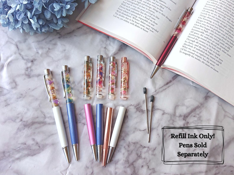 Peut inclure: Un ensemble de huit stylos avec des fleurs color&eacute;es et des paillettes enferm&eacute;es dans de la r&eacute;sine transparente. Les stylos ont des corps de diff&eacute;rentes couleurs, notamment blanc, bleu, rose et or rose. Le texte "Recharge d'encre uniquement ! Stylos vendus s&eacute;par&eacute;ment" est visible sur l'image.