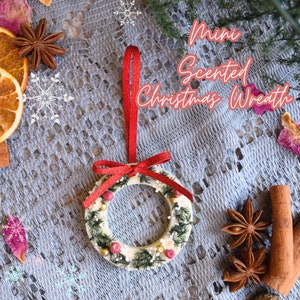 May include: A white, mini Christmas wreath with a red ribbon and green foliage. The wreath is decorated with small white flowers, red berries, and gold beads. The wreath is hanging from a red ribbon and is on a light blue background. The text "Mini Scented Christmas Wreath" is written in pink letters on the top of the image.