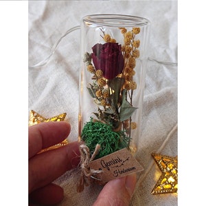 May include: A glass tube filled with dried flowers, including a red rose, and green moss. A small brown tag is attached to the tube with the text "Gemini Herbarium".