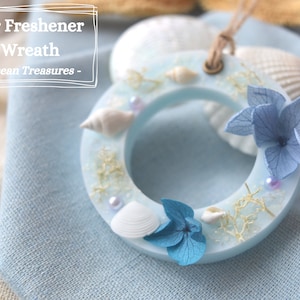 May include: A light blue wax air freshener wreath with a seashell, dried flowers, and pearl accents. The wreath is labelled "Air Freshener Wreath - Ocean Treasures -"