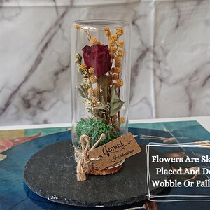 May include: A glass tube filled with a dried red rose and yellow flowers, with a cork stopper and a brown tag that says "Gemini Herbarium". The tube is sitting on a black slate coaster.