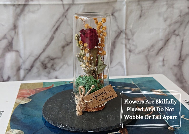 May include: A glass tube filled with a dried red rose and yellow flowers, with a cork stopper and a brown tag that says "Gemini Herbarium". The tube is sitting on a black slate coaster.
