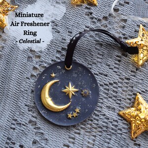 May include: A black and gold celestial-themed air freshener ring with a crescent moon, stars, and a sun. The ring is hanging from a black ribbon.