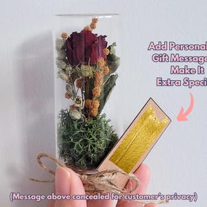 May include: A glass tube filled with a preserved red rose and moss, with a cork stopper and a small tag attached. The tag has a gold background and is blank, allowing for a personalized message.