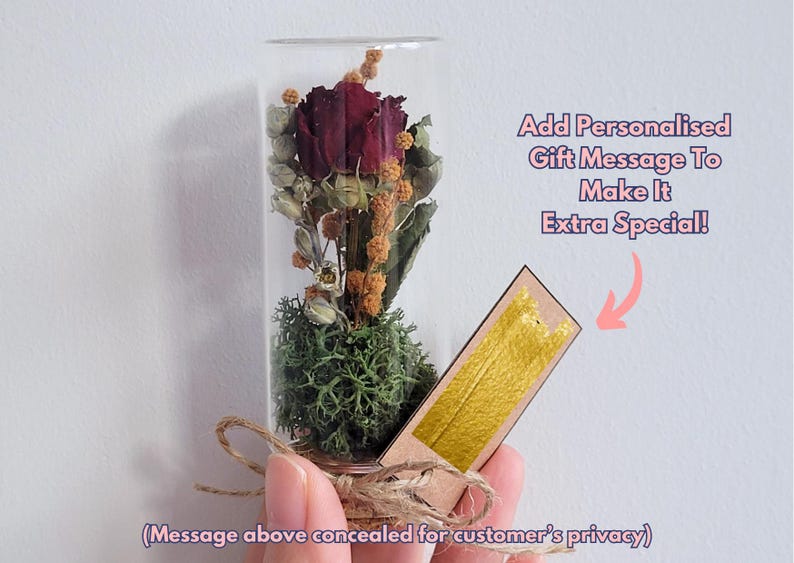 May include: A glass tube filled with a preserved red rose and moss, with a cork stopper and a small tag attached. The tag has a gold background and is blank, allowing for a personalized message.