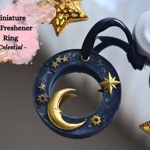 May include: A navy blue air freshener ring with a gold crescent moon, stars, and a sun design. The ring is hanging from a black ribbon and has the text "Celestial" on it.